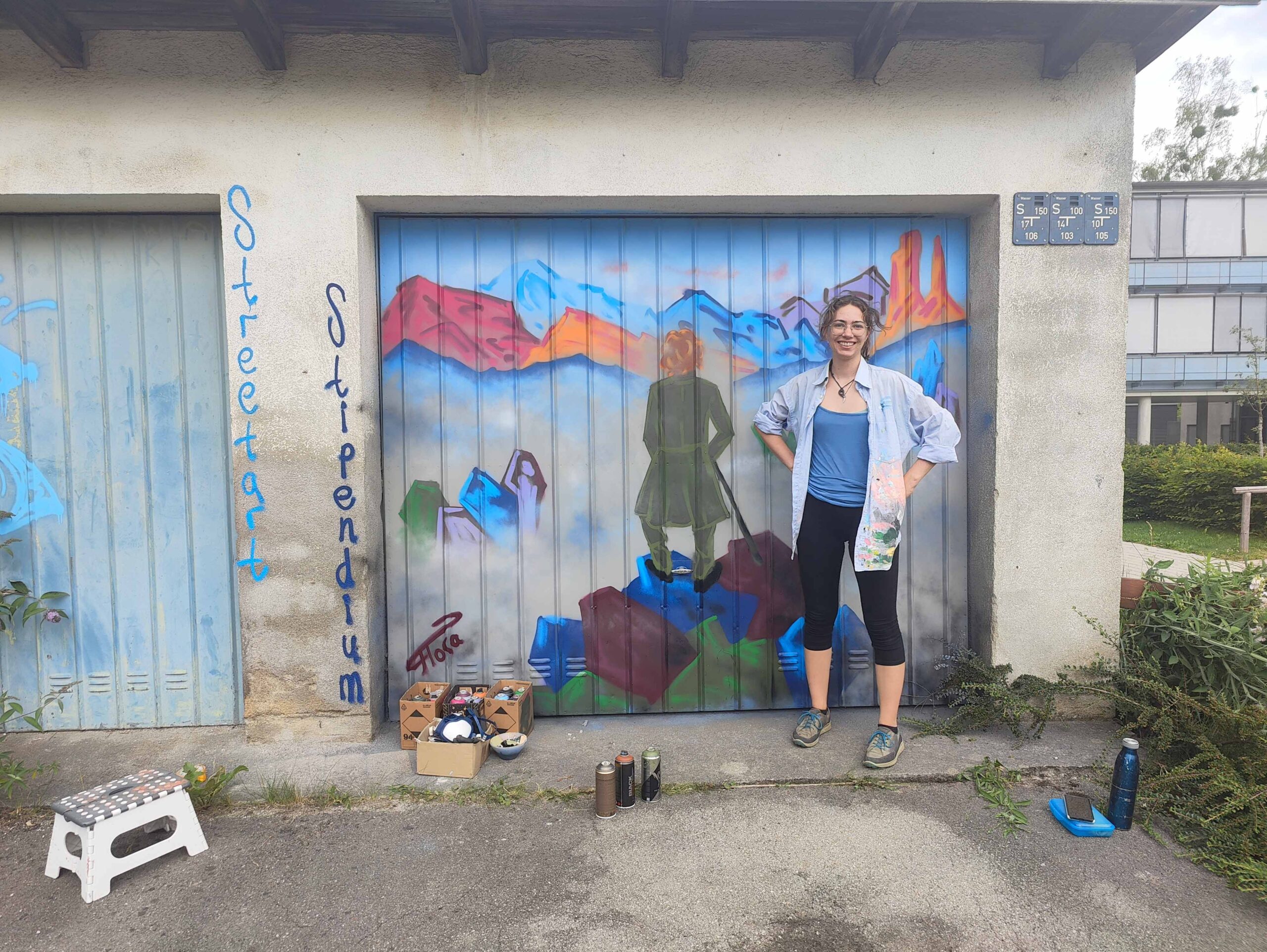 U27 Streetart Stipendium Lansberg 2024: Emma vor ihrem gestalteten Garagentor in Schondorf am Ammersee. Das Werk zeigt eine Interpretation vom Wanderer im Nebelmeer von Caspar David Friedrich