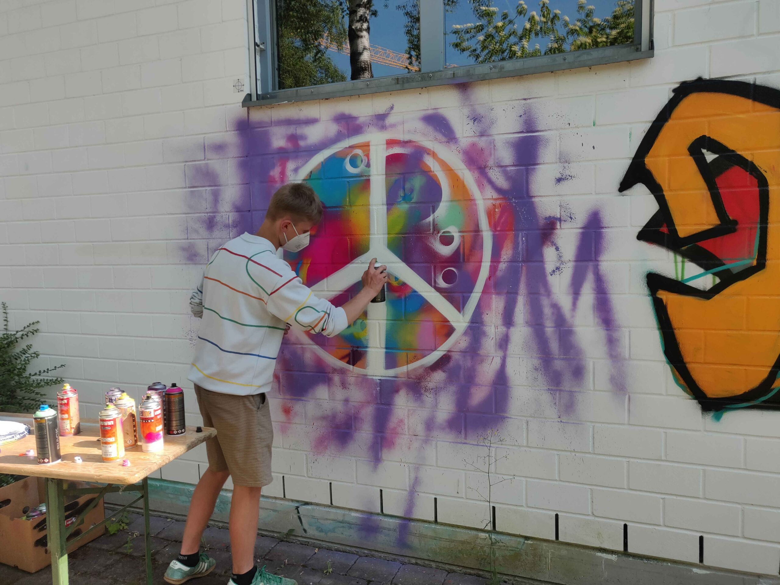 Ein Jugendlicher sprüht während eines Streetart und Graffiti Workshops ein Peace Zeichen an die Wand mit einem bunten Hintergrund. Urbane kunst und Kultur by Erwa.One