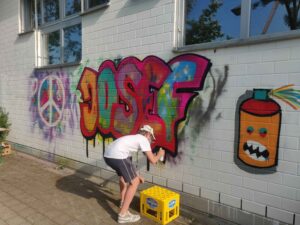 Ein Jugendlicher sprüht während eines Streetart und Graffiti Workshops ein "Josef"-Graffiti an die Wand mit bunten Fill-Ins. Jugendarbeit by Erwa.One