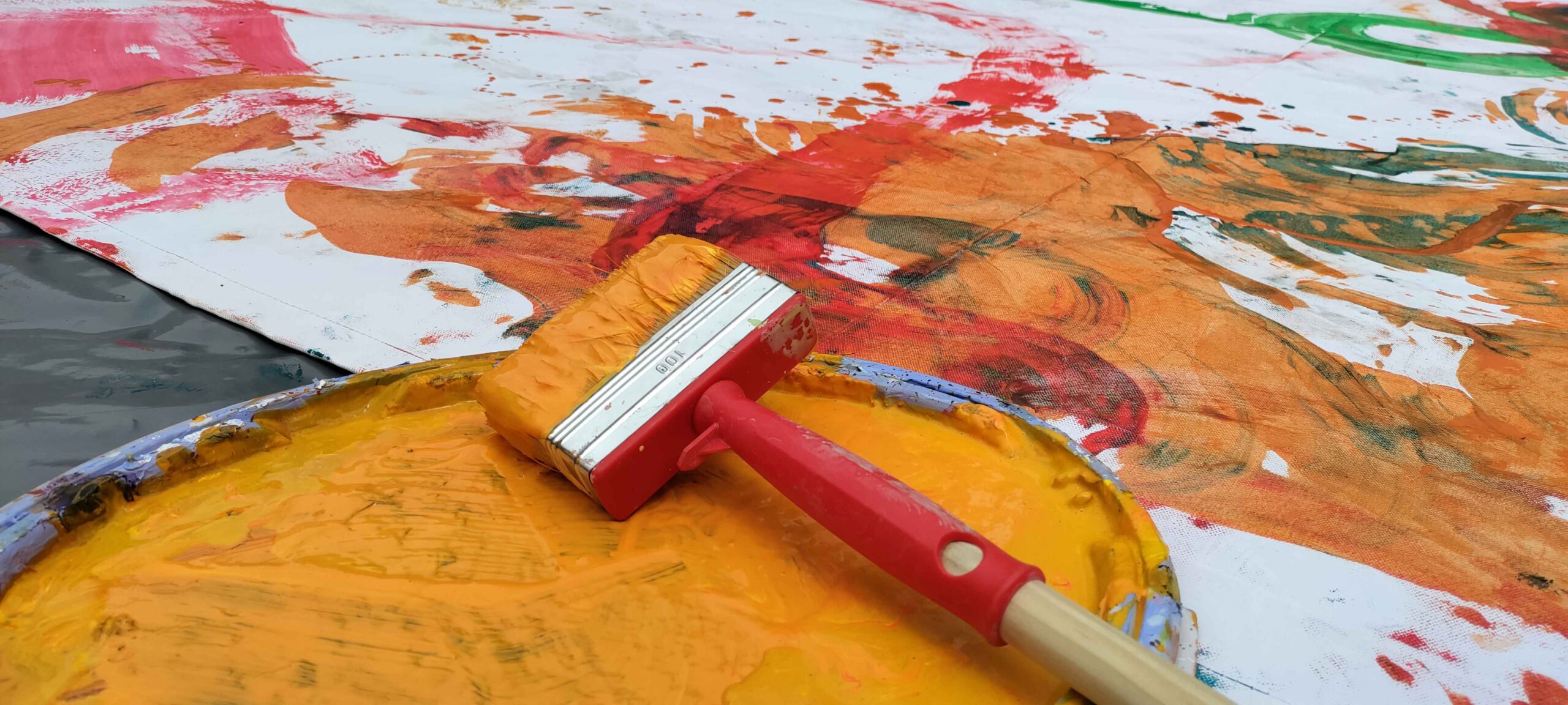 gelber Pinsel auf Deckel im Vordergrund. Dahinter Plane mit bunten Farbklecksern