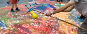 Person zieht an Schnur und verspritzt durch eine Vorrichtung Farbe auf eine Plane am Boden. Workshop mit anderen Personen im Hintergrund und einem bunt bemalten Untergrund in Form einer ausgebreiteten LKW Plane