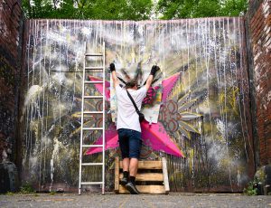 Streetart-Künstler Erwa.One bei der Arbeit. Die Arme gestreckt, halten eine große Schablone, welche er gegen ein 3 Meter hohes Tor drückt. Eine Leiter steht neben dem Streetartist. Köln-Ehrenfeld Kolbhalle Artist Community