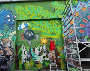 Der Künstler ErwaOne in einem roten Pulli bei der Arbeit während der Graffiti Jam Bavarian Safari in Landsberg. Streetartmotiv O-Bomb aus Mario Kartb auf grünem Hintergrund. Gemeinschaftswerk