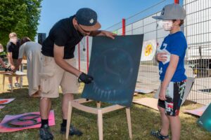 StreetartKünstler Erwa.One während eines B2B Firmenevents mit einer Streetart Station, 2022. Der Streetartist erklärt einem Jungen wie man auf einen Karton sprüht.