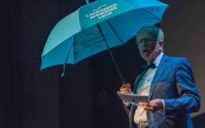 Kulturreferent Axel Flörke 2022 während der Preisvergabe des Kulturförderpreises in Landsberg am Lech auf der Bühne des Stadttheaters mit einem blauen Regenschirm
