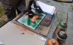 Eine Person entfernt eine frisch besprühte Schablone von einem papier. Sprühdosen auf dem Tisch, Bunte Flecken auf der Unterlage