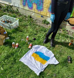 Auf einer Wiese liegt ein Tshirt mit Ukraine Farbe Gelb Blau und Sprühdosen daneben. Dahinter ein Mädchen mit schwarzen Schuhen und eine bunt bemalte Graffiti Wand