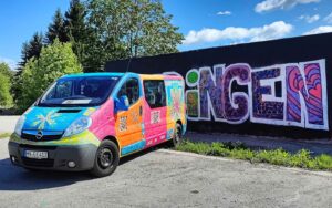 Neue Ornamente-Gestaltung von Erwa.One auf dem Juze-on-Tour-Mobil vom Kreisjugendring Unterallgäu. Der ganze Bus ist von der Entfernung zu sehen, sowie die Graffiti-Wand für den Einführungsworkshop im Hintergrund. Darauf steht Ettringen. Der Bus wurde mit filigranen Streetart-Schablonen besprüht, Die Schablonen erinnern an Masken und sind in bunten Geometrien angeordnet.