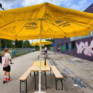 Location Sprühworkshop und Spray my day Festival Herrsching während dem sommerlichen Wochenende, Bierbänke und Sonnenschirme wurden aufgestellt und ein Mädchen fährt auf ihrem Roller vorbei