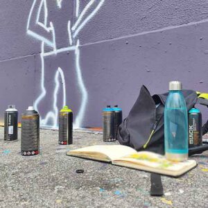 Ambiente Bild, Sketchbook mit Sprühdosen vor einer Wand mit angefangenem Graffiti-Charakter, Wasserflasche und Rucksack daneben