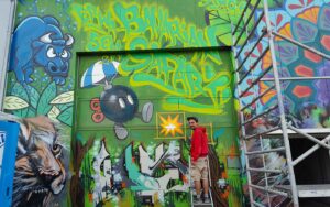 Bavarian Safari Graffiti Jam Landsberg 2023 ErwaOne vor bunten Fassade Gesamtkunstwerk, sprüht letzte Details O-Bomb und illustrative Explosionsfetzen im MarioKart Style