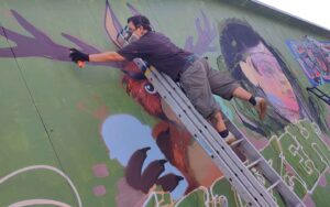Bavarian Safari Graffiti Jam Landsberg 2023 LouZeh Hip Hop Künstler bei der Arbeit, balanciert mit einem Fuß auf einer Leiter um das Geweih von seinem Humpoltinger-Fantasiewesen zu sprühen