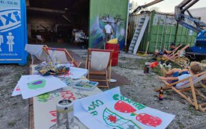 Bavarian Safari Graffiti Jam Landsberg 2023 Veranstaltungswochenende Malstation mit Kinderbildern, Menschen auf Liegestühlen, Künstler mal Löwe, rechts Hebebühne, offenes Tor zur Gewerbehalle