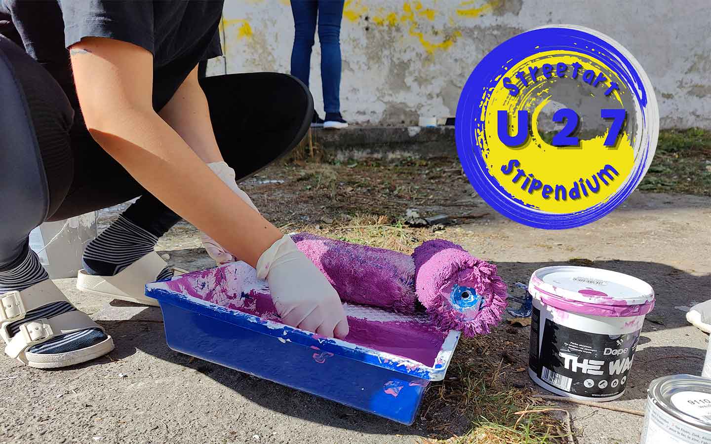 U27 Streetart Stipendium Landsberg am Lech Jugendarbeit, Junge Künstler mit Farbwanne und Farbroller, rechts das Logo des Stipendiums in blau gelber Farbe Open-Call Ausschreibung