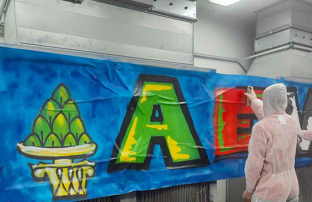 Graffiti-Sprühworkshop in der Handwerkskammer Schwaben mit den Nachwuchsspielern der Augsburger Panther. Ein Junge in Schutzkleidung sprüht in einem Lackierraum ein rotes E, als Buchstaben auf einen blauen Hintergrund.
