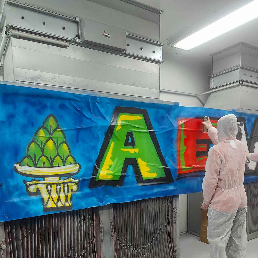 Graffiti-Sprühworkshop in der Handwerkskammer Schwaben mit den Nachwuchsspielern der Augsburger Panther. Ein Junge in Schutzkleidung sprüht in einem Lackierraum ein rotes E, als Buchstaben auf einen blauen Hintergrund.