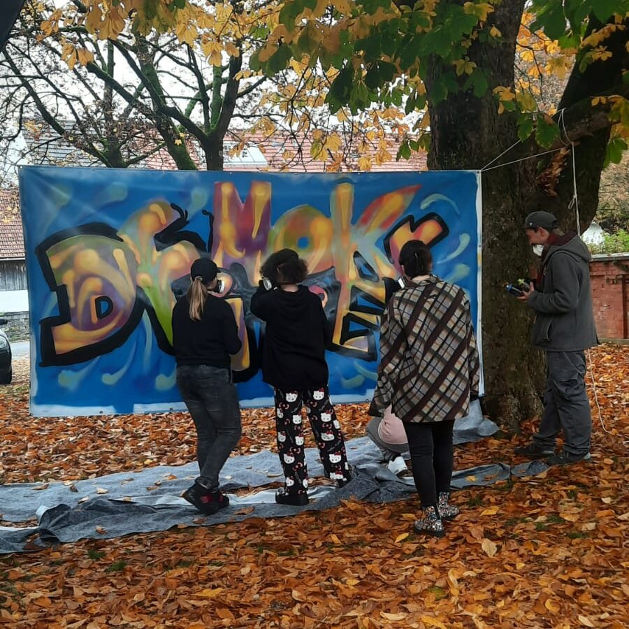 Demokratiebildung Bannergestaltung mit dem Jugendparlament von Ettringen Streetart Kunst Projekt mit Erwa.One und dem Kreisjugendring Unterallgäu Einführungssprühkurs Graffiti mit dem Wort "Demokratie" in bunten Farben, kunstpädagogische Vermittlung