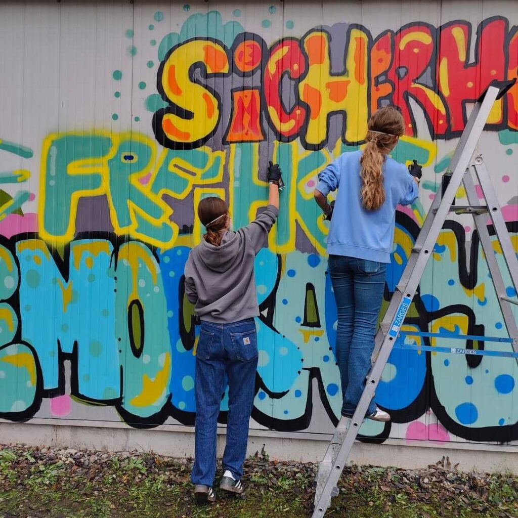 Foto der "Words for Democracy" Kunstprojektes mit dem Kreisjugendring Landsberg und dem Streetart-Künstler Erwa.One im Jugendhaus Lechside Lounge in Kaufering, Bayern. zwei junge Sprüherinnen gestalten ein blau-gelbes Graffiti mit einer Leiter und vielen Sprühdosen. Words for Democracy Kunstprojekt