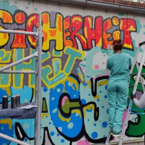Words for Democracy Kunstprojekt mit ErwaOne in Kaufering. Workshop Demokratiebildung und Wandgestaltung mit Schulklasse. Ein Mädchen steht auf einer Leiter und besprüht die Wandfläche. In Zusammenarbeit mit dem Kreisjugendring Landsberg