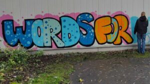 Ein Mädchen sprüht während einem Graffiti-Workshop ein Bild an die Wand. Links ist der Schriftzug Words for... zu lesen. Kunstvermittlung durch Erwa.One urbane Kunst und Streetart