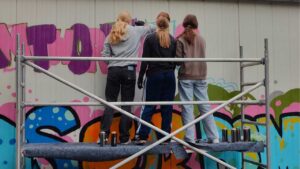 Foto der "Words for Democracy" Kunstprojektes mit dem Kreisjugendring Landsberg und dem Streetart-Künstler Erwa.One im Jugendhaus Lechside Lounge in Kaufering, Bayern. Drei junge Sprüherinnen gestalten ein pink-violettes Graffiti und stehen auf einem Gerüst und vielen Sprühdosen