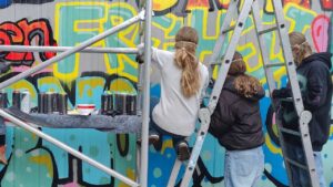 Foto der "Words for Democracy" Kunstprojektes mit dem Kreisjugendring Landsberg und dem Streetart-Künstler Erwa.One im Jugendhaus Lechside Lounge in Kaufering, Bayern. Drei junge Sprüherinnen gestalten ein blau-gelbes Graffiti mit einer Leiter und einem Gerüst und vielen Sprühdosen