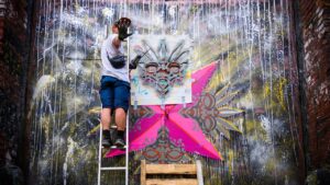Urban Art Künstler ErwaOne gestaltet ein Streetart-Werk mit Schablonen am Tor der Kolbhalle Helmholtzstrasse Köln-Ehrenfeld. Er zeigt eine Handfläche in die Kamera und steht auf einer Leiter vor seinem halb fertigen Werk