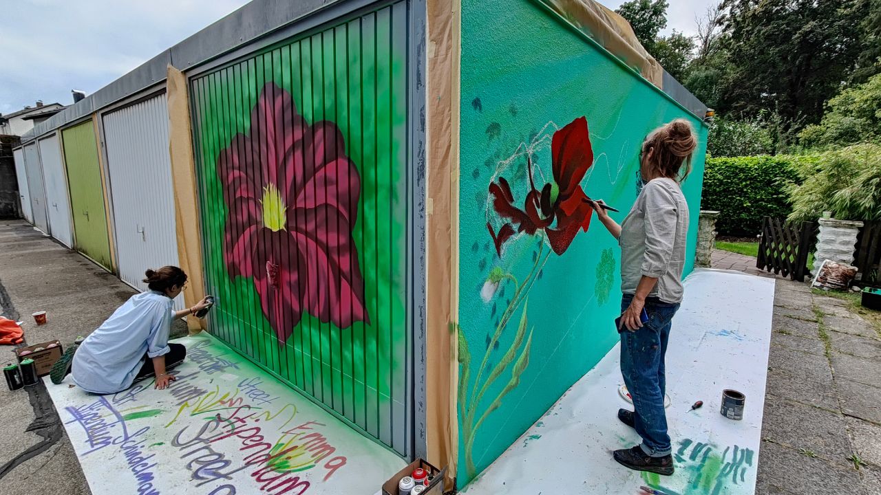 Lucce x Erwa.One und das U27 Streetart-Stipendium. Stipenidatin Emma sprüht links eine Blume auf ein Garagentor im Osten von Landsberg. Rechts malt die Künstlerin Lucceein Streetart-Werk an eine Garagenwand