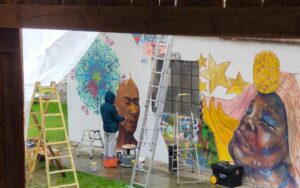 Streetart-Begegnung zwischen den Kulturen mit Lucce aus Toulouse und Erwa.One aus Landsberg. Wandgestaltung am Unser Theater Schwabhausen im Regen trotz Sommer mit einem Portrait von Frida Kahlo und dem Schablonen-Stil der neuen Ornamente. Sprühdose und Pinsel Kombination, interkulturelle Zusammenarbeit