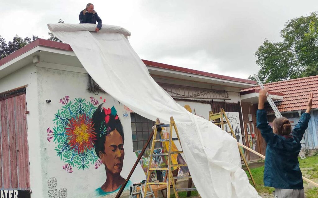 Streetart-Begegnung zwischen den Kulturen mit Lucce aus Toulouse und Erwa.One aus Landsberg. Helfer spannen eine Behelfsplane als regenschutz aus