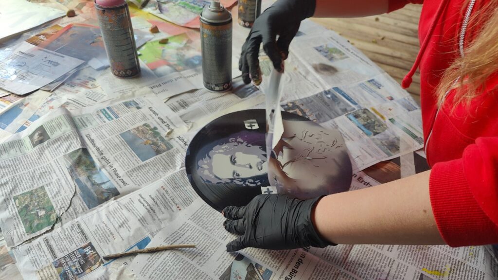 Sprüh-Workshop Urbane Kunst Methodik von ErwaOne an der Montessori-Schule von Kaufering. Ein Mädchen zieht eine Schablone von einer Schallplatte ab. Der Betrachter kann ein Protrait von Schiller erahnen. dreilagige Schablone Teil des Workshops zu Stencil-Kunst (wie Banksy, C215, ...)