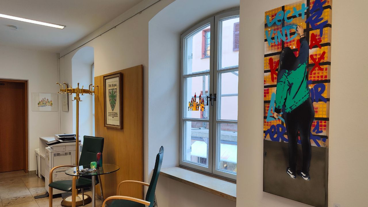 Viva la Buntness Graffiti & Streetart Ausstellung im Bürgerbüro von Aichach-Friedberg 2023. Lechkind-Leinwand Werk von ErwaOne. Stühle stehen im Raum. Fenster gibt den Blick auf die Straße und die Altstadt