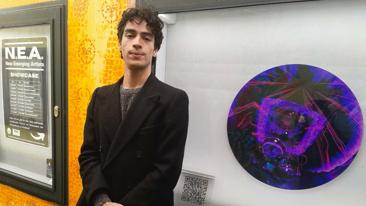 N.E.A. - New emerging artists Ausstellungsreihe in der Zederpassage von Landsberg am lech zeigt neue künstlerische Positionen. Der kolumbianische Künstler Juan Rubiano welcher in München wohnt steht neben dem Schaufenster mit seinem KusntwerkVision und schaut in die Kamera. Die Passage hinter ihm ist mit streetartmotiven gestaltet.