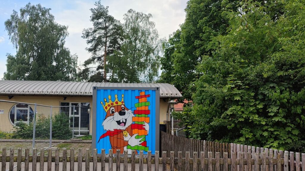 Streetart Gestaltung einer Container-Fläche mit dem Motiv eines Biber welcher einen Turm aus Holzklötzen in der Hand hält und lächelt. Ort Kindergarten Biberburg in Scheuring bei Landsberg am Lech. Auftragsarbeit Kunst im öffentlichen Raum by Erwa.One