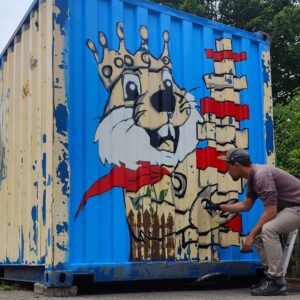 Streetart Gestaltung einer Container-Fläche mit dem Motiv eines Biber welcher einen Turm aus Holzklötzen in der Hand hält und lächelt. Ort Kindergarten Biberburg in Scheuring bei Landsberg am Lech. Auftragsarbeit Kunst im öffentlichen Raum by Erwa.One Das Bild ist noch nicht fertig und der Künstler (rechts im Bild) sprüht am unteren Teil des Bildes.