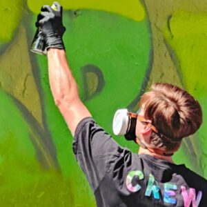 Ein Mann sprüht ein Graffiti in grünen Farbtönen. Auf seinem T-Shirt Kleidungsstück steht CREW