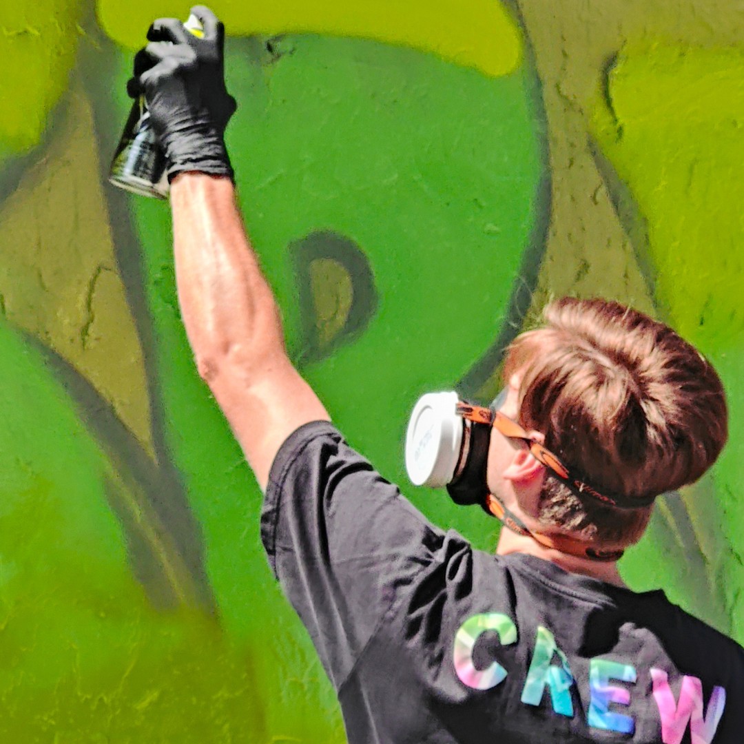 Ein Mann sprüht ein Graffiti in grünen Farbtönen. Auf seinem T-Shirt Kleidungsstück steht CREW