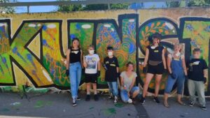 Intergenerationeller Streetart- und Graffiti Workshop mit ErwaOne in Landsberg am Lech an der legalen Sprühfläche der Stadt. Gruppenfoto der Teilnehmer vor dem partizipativen Kunstwerk, welches Teil der Einführung war. Das Wort Kunst ist im Hintergrund zu lesen