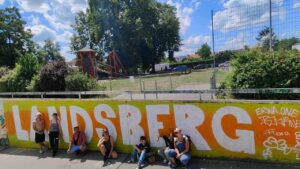 Intergenerationeller Streetart- und Graffiti Workshop mit ErwaOne in Landsberg am Lech an der legalen Sprühfläche der Stadt. Gruppenfoto vor dem partizipativen Kunstwerk Landsberg Graffito als Schriftzug
