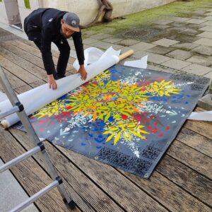 Kunstkanal Augsburg Installation von Urban Art Streetart Künstler ErwaOne am Lechkanal in der Altstadt. Sommer. Der Künstler packt eine bunt gestaltete Plane von einer Rolle aus. Eine Leiter ist links im Bild zu sehen.
