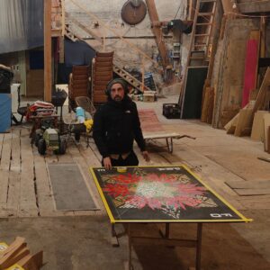 Der Künstler Erwa.One aus dem Streetart / urbane Kunst gestaltet ein Holzpanel in einer Scheune, welches an ein Klavier (links im Bild) angebracht wird). Er trägt Kopfhörer und eine Mütze, schaut in die Kamera.