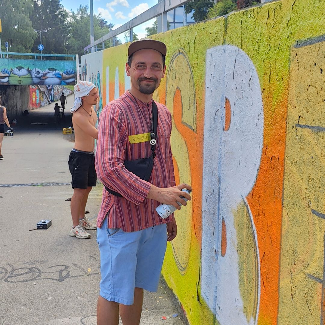 Intergenerationeller Streetart- und Graffiti Workshop mit ErwaOne in Landsberg am Lech an der legalen Sprühfläche der Stadt. Der Künstler trägt ein rotes Leinenhemd und lächelt in die Kamera. Er hält eine Sprühdose in der Hand.