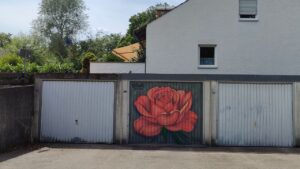 Bunt gestaltetes Garagentor in Landsberg am Lech, Oberbayern Deutschland. Streetart-Motiv Rose in rosé-Farbtönen. Ein Haus im Hintergrund mit Terasse. Sommer