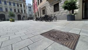 Print the City mit ErwaOne und Kunstsammlungen und Museen Augsburg am Fuggerplatz neben dem Maximiliansmuseum. Ein Gulli-Deckel mit dem Motiv der Zirbelnuss ist zu sehen.