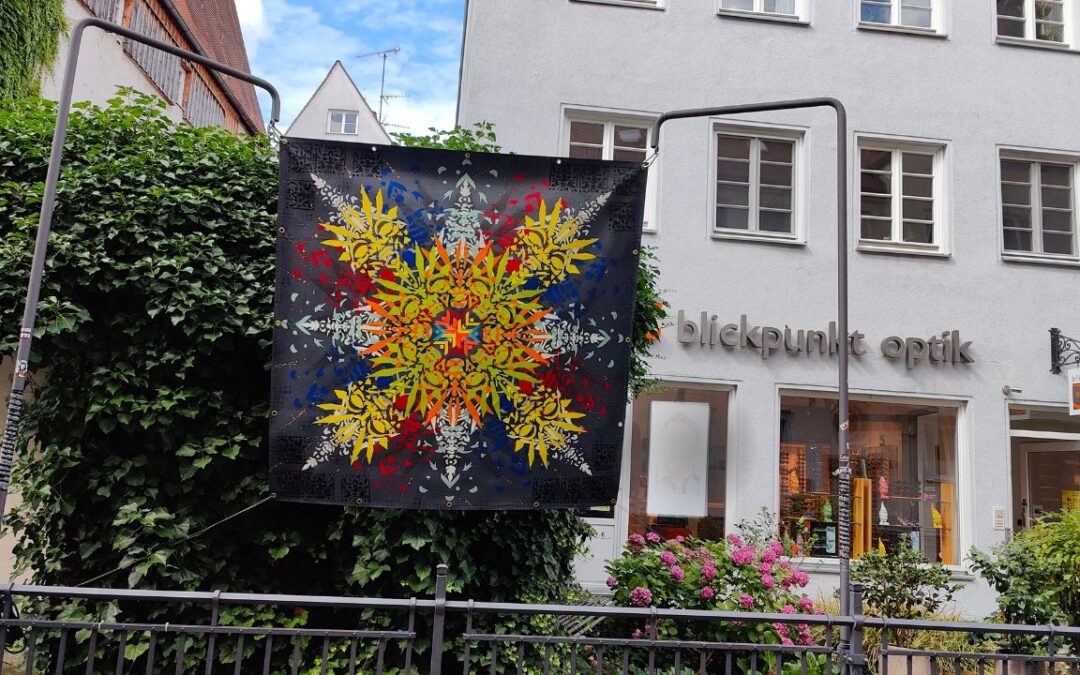 Kunstkanal Augsburg Installation von Urban Art Streetart Künstler ErwaOne am Lechkanal in der Altstadt. Sommer. Eine bunt gestaltete Plane hängt von zwei Ständern über einem Wasserkanal.