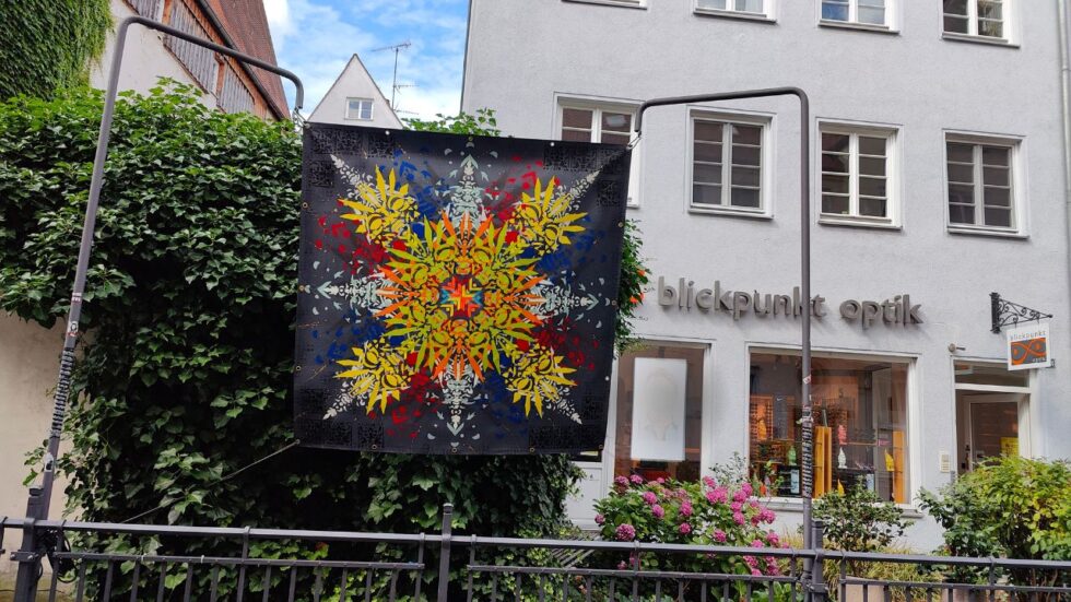 Kunstkanal Augsburg Installation von Urban Art Streetart Künstler ErwaOne am Lechkanal in der Altstadt. Sommer. Eine bunt gestaltete Plane hängt von zwei Ständern über einem Wasserkanal.