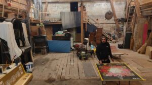 Der Künstler Erwa.One aus dem Streetart / urbane Kunst gestaltet ein Holzpanel in einer Scheune, welches an ein Klavier (links im Bild) angebracht wird). Er trägt Kopfhörer und eine Mütze, schaut in die Kamera.