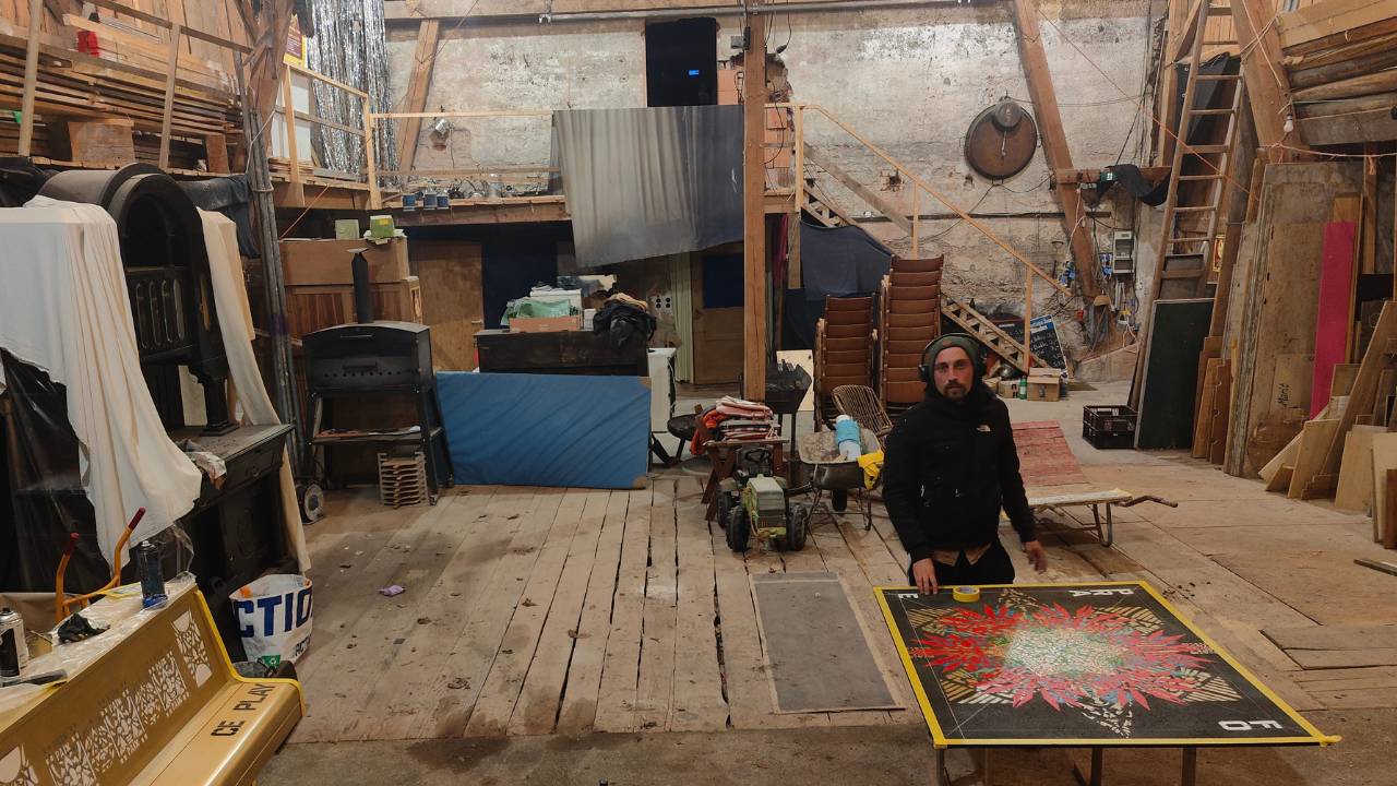 Der Künstler Erwa.One aus dem Streetart / urbane Kunst gestaltet ein Holzpanel in einer Scheune, welches an ein Klavier (links im Bild) angebracht wird). Er trägt Kopfhörer und eine Mütze, schaut in die Kamera.