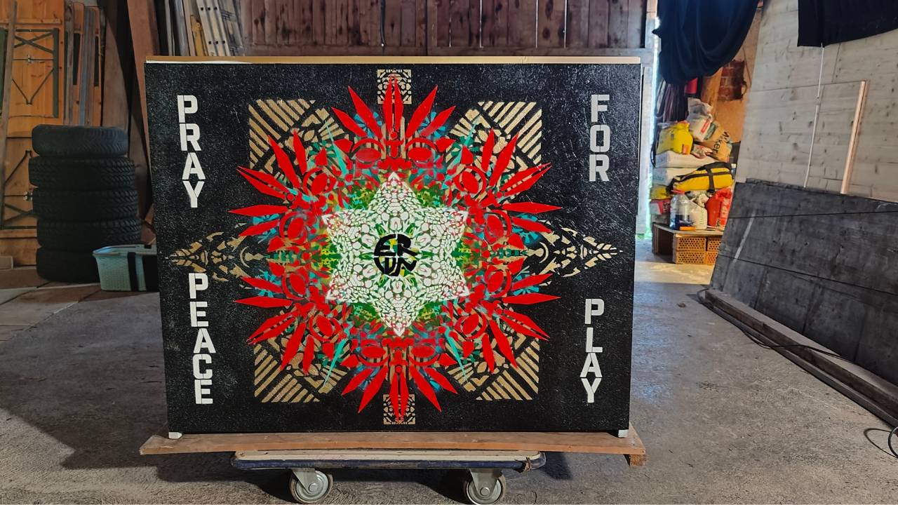Bunt gestaltetes Klavier design by Streetart Künstler Erwa.One. Das Instrument steht in einer Scheune, welche als provisorisches Atelier zur Gestaltung diente. Die Rückseite ist zu sehen mit Ornamenten auf schwarzem Hintergrund und dem Satz Pray for Peace Play. Das Klavier wird für die 9.Play Me Edition in Augsburg im öffentlichen Raum ausgestellt und darf von Passanten bespielt werden.