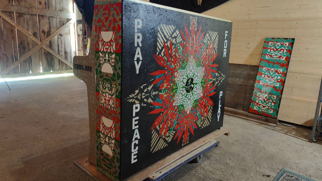 Bunt gestaltetes Klavier design by Streetart Künstler Erwa.One. Das Instrument steht in einer Scheune, welche als provisorisches Atelier zur Gestaltung diente. Die Rückseite ist zu sehen mit Ornamenten auf schwarzem Hintergrund und dem Satz Pray for Peace Play. Das Klavier wird für die 9.Play Me Edition in Augsburg im öffentlichen Raum ausgestellt und darf von Passanten bespielt werden.